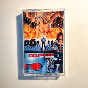 May include: A transparent cassette tape with the cover art for the "AD. Police" soundtrack. The artwork shows a futuristic city with explosions, figures in armour, and the text "AD. POLICE" in a bold font.