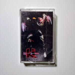 Op de afbeelding: Een vintage cassettebandje met het spel "Killer Instinct". De cover toont een donkere, actiegerichte illustratie van een personage met gloeiende paarse klauwen en het rode logo van het spel. De cassette zit in een transparante plastic hoes.