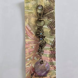 Puede incluir: Un llavero de metal color bronce con cierre de mosquetón, un dije de flor, un dije de corazón y un colgante redondo de color morado iridiscente. El llavero está sujeto a una tarjeta rectangular con estampado floral y el texto handmade with love by Yippie Gypsy.