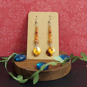 May include: A pair of dangle earrings with silver hooks. The earrings feature a combination of orange beads, small stones, and gold-colored circular pendants. The earrings are displayed on a brown card against a red floral background.