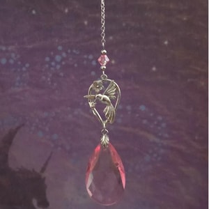 May include: A silver-toned hummingbird suncatcher with a pink teardrop-shaped crystal. The suncatcher features a hummingbird design with a flower and a small pink crystal bead. The background is a purple and blue gradient.
