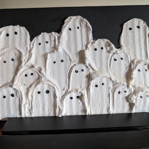 May include: A Halloween-themed artwork featuring a textured, white ghost design on a black canvas. The ghosts have black dot eyes and are arranged in a layered, clustered composition. The artwork is displayed on a black stand.