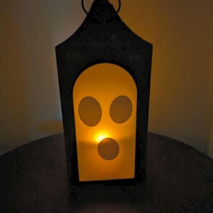 May include: Black metal lantern with a spooky face design. The lantern has a handle at the top and a glowing orange light inside, creating a Halloween or spooky theme. The face has three circular eye holes and a circular mouth hole.