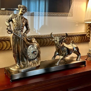 May include: A gold-toned tabletop clock featuring a bullfighter figurine holding a cape and a charging bull figurine. The clock face is visible with Roman numerals and black hands.