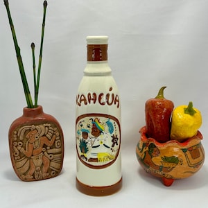 May include: A white ceramic bottle with a brown label that reads "Kahlua". The bottle features a colourful design with a person in a hat and a red, white and blue colour scheme. The bottle is sitting on a white surface next to a brown ceramic vase with a green plant and a small ceramic bowl with red and yellow peppers.