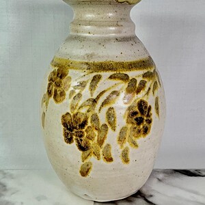 May include: Cream-colored ceramic vase with a wide body and narrow neck. The vase features a band of brown floral designs around the middle and top. The rim and the band of floral designs are a darker brown color.