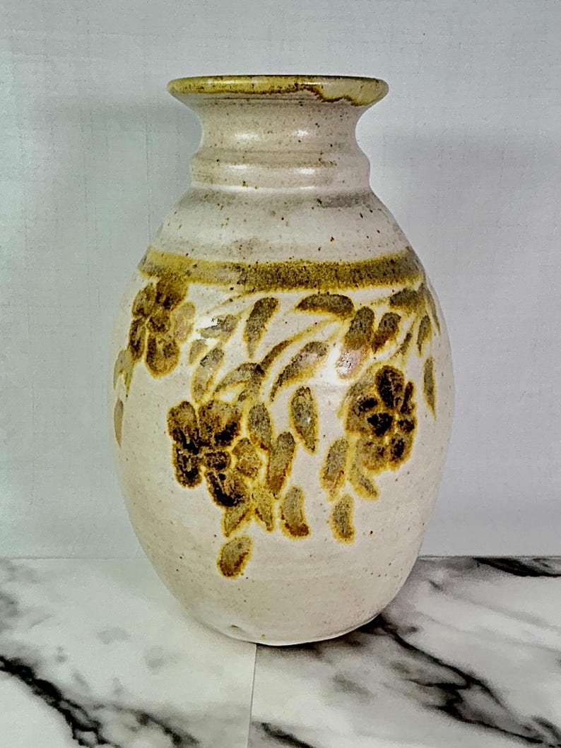 May include: Cream-colored ceramic vase with a wide body and narrow neck. The vase features a band of brown floral designs around the middle and top. The rim and the band of floral designs are a darker brown color.
