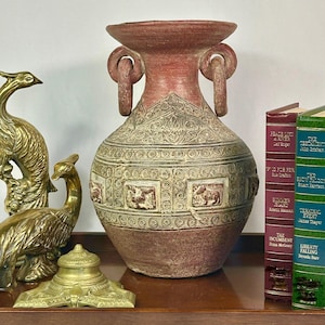 May include: A large, reddish-brown ceramic vase with ornate carvings and loop handles. The vase features a detailed design with animal figures and geometric patterns. Decorative brass peacock figurines and books are also visible.