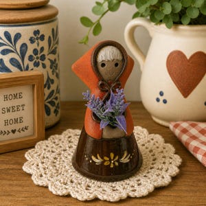 May include: A ceramic figurine of a person in a hooded cloak holding purple flowers. The figurine has a brown skirt with yellow floral accents. The background includes a decorative jar, a framed sign that reads "HOME SWEET HOME", and a crocheted doily.