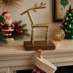 May include: A gold-coloured metal reindeer Christmas stocking holder on a wooden base. The reindeer has a simple, geometric design with antlers and is placed on a white mantel with other holiday decorations.
