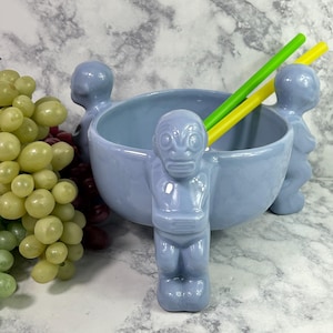 May include: A light blue ceramic tiki bowl with three tiki figures attached. The bowl has a wide opening and contains two straws, one yellow and one green. Green and red grapes are in the background.