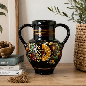 May include: A black ceramic vase with two handles, decorated with vibrant floral patterns in yellow, red, and green. The vase has a glossy finish and is displayed on a wooden surface. A decorative home decor item.