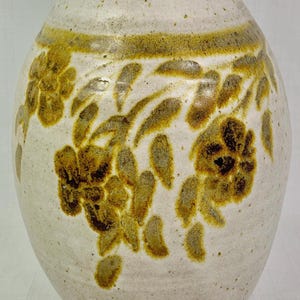 May include: Cream-colored ceramic vase with a rounded body and a narrow neck. The vase features a band of brown and yellow floral designs around the middle. The flowers and leaves are hand-painted, adding a rustic touch to the decorative piece.