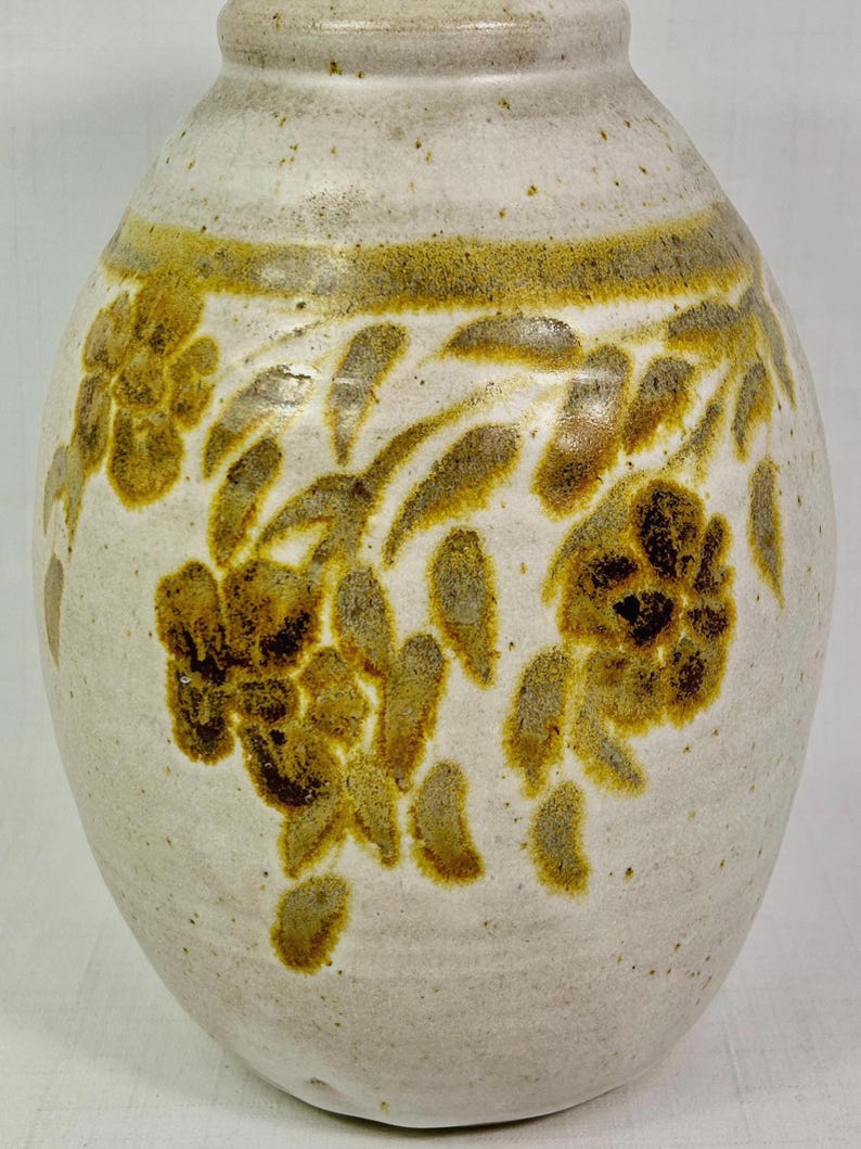 May include: Cream-colored ceramic vase with a rounded body and a narrow neck. The vase features a band of brown and yellow floral designs around the middle. The flowers and leaves are hand-painted, adding a rustic touch to the decorative piece.