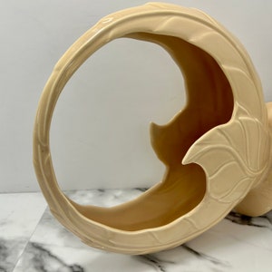 May include: A cream-colored ceramic basket with a curved handle and leaf-like details. The basket has a crescent shape with a wide opening and a textured surface. The design is a decorative piece for home decor.