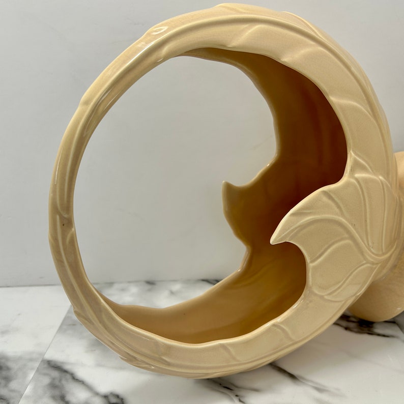 May include: A cream-colored ceramic basket with a curved handle and leaf-like details. The basket has a crescent shape with a wide opening and a textured surface. The design is a decorative piece for home decor.