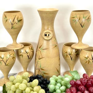 May include: A tan ceramic drinkware set featuring a tall pitcher with a button detail and six glasses. Each piece is decorated with a bamboo design in black and green. The set is displayed with artificial grapes in various colours.
