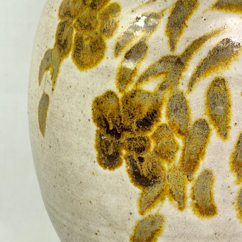 May include: A ceramic vase with a cream-colored base and a floral pattern in shades of brown and yellow. The flowers and leaves are painted in a textured style, creating a rustic aesthetic.