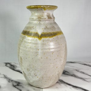 May include: A handcrafted ceramic vase with a speckled, off-white glaze. The vase features a flared rim and a subtle, curved design with a band of olive green and yellow around the middle. The vase has a rounded body and a smooth, textured surface.