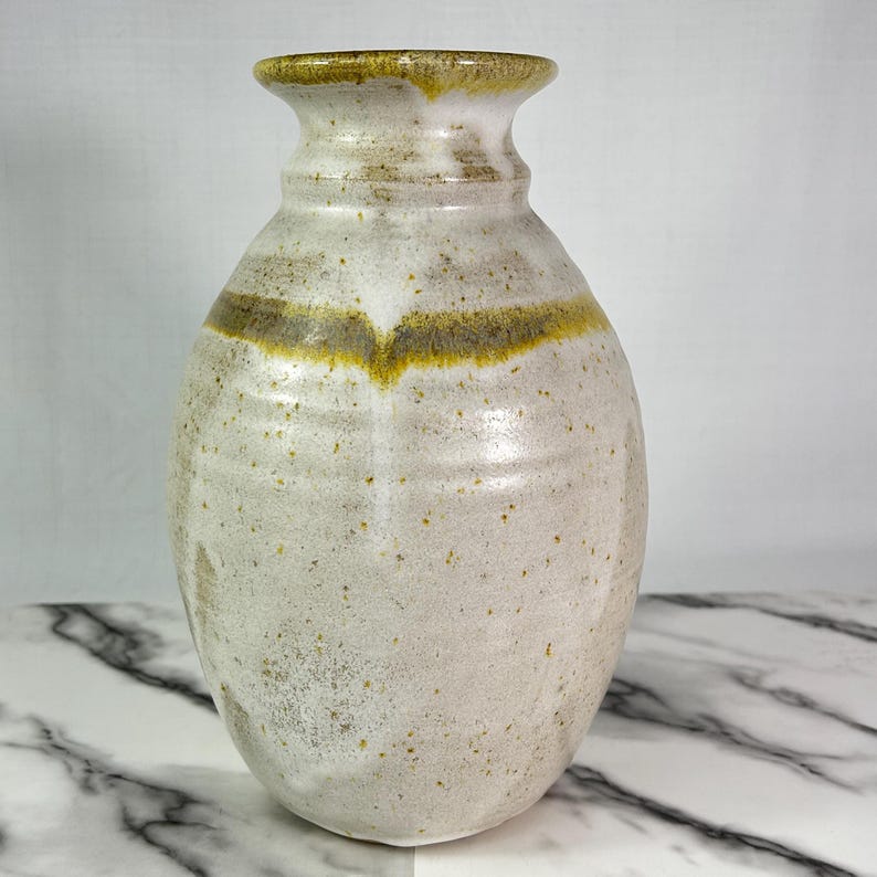 May include: A handcrafted ceramic vase with a speckled, off-white glaze. The vase features a flared rim and a subtle, curved design with a band of olive green and yellow around the middle. The vase has a rounded body and a smooth, textured surface.
