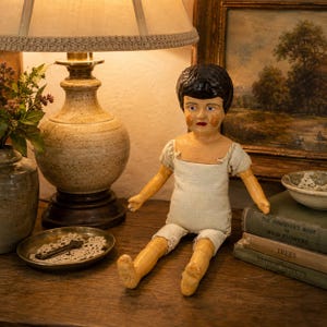 May include: A vintage doll with painted features, black hair, and a white outfit sits on a wooden surface. The doll is next to a lamp with a beige lampshade and a decorative base. A small dish and books are also visible.