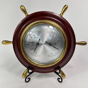 May include: A decorative ship's wheel-shaped hygrometer and thermometer. The wooden frame is a dark reddish-brown with gold-coloured accents. The face displays temperature in Fahrenheit and humidity levels. The base is black metal.
