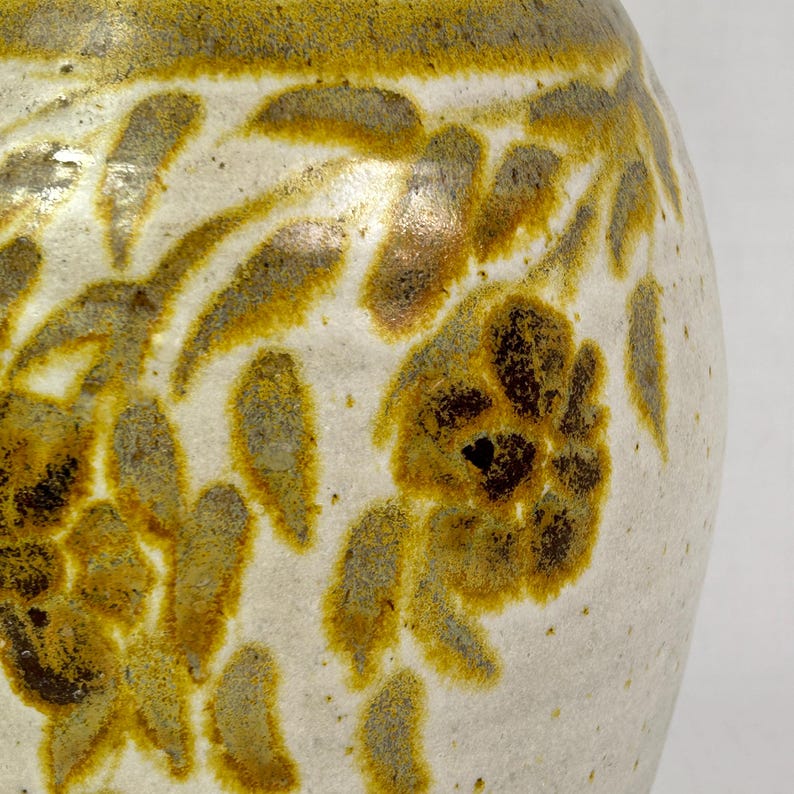 May include: A close-up of a ceramic vase with a white base and a floral design in shades of yellow and brown. The flowers and leaves are painted in a stylized, organic pattern, with a textured, speckled finish.
