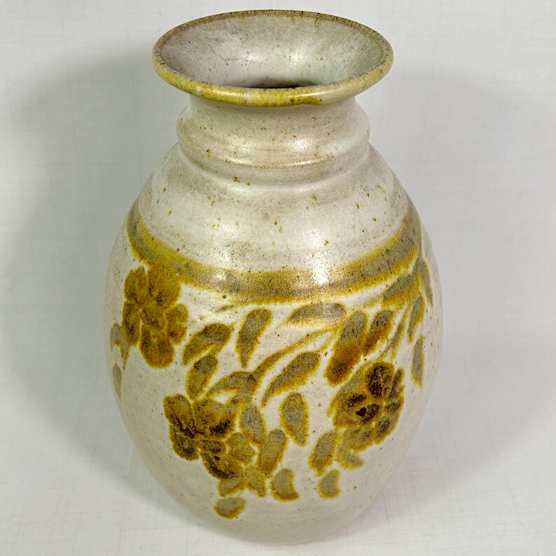 May include: A ceramic vase with a wide, rounded body and a narrow neck. The vase is primarily off-white with a band of gold around the middle and floral designs in gold. The opening is 4 inches in diameter.