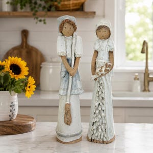 May include: Two ceramic figurines of women in long dresses and bonnets. One holds a broom, the other a bundle of flowers. The figures are primarily white with blue and brown accents. The figurines are standing on a marble countertop.