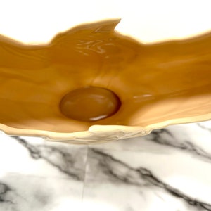 May include: A beige ceramic decorative bowl with a glossy finish. The bowl has a unique, organic shape with a rounded base and a raised, petal-like rim. The interior is a darker shade of beige than the exterior.