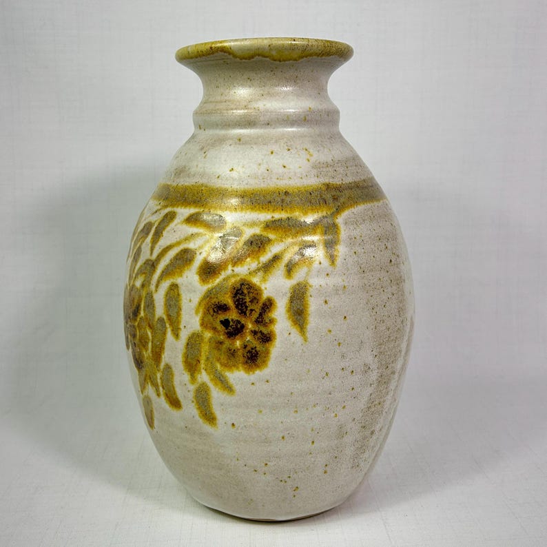 May include: A handcrafted ceramic vase with a cream-colored base and a brown floral design. The vase has a rounded body and a narrow neck. The floral pattern is repeated around the vase. The vase is approximately 10 inches tall.
