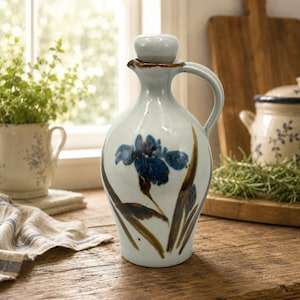 May include: A ceramic oil cruet with a glossy off-white glaze, featuring a hand-painted blue iris flower with green and brown leaves. The vessel has a rounded body, a curved handle, a narrow neck, and a matching removable spherical stopper.