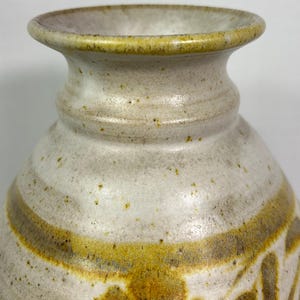 May include: A handcrafted ceramic vase with a speckled, off-white finish. The vase features a wide, decorative band in shades of gold and brown, with floral and leaf motifs. The rim and base have a subtle yellow-green hue, adding to its rustic charm.