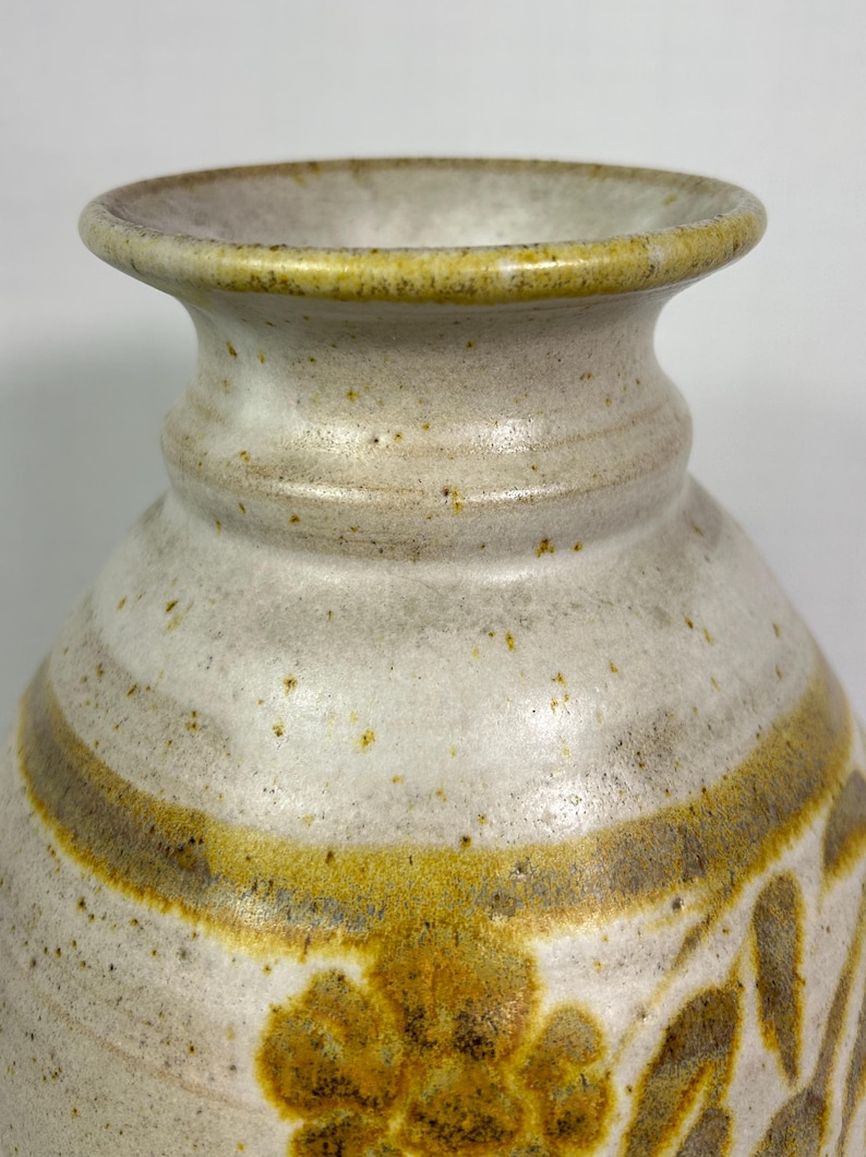 May include: A handcrafted ceramic vase with a speckled, off-white finish. The vase features a wide, decorative band in shades of gold and brown, with floral and leaf motifs. The rim and base have a subtle yellow-green hue, adding to its rustic charm.