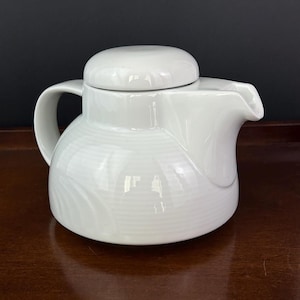 May include: A white ceramic teapot with a lid, handle, and spout. The teapot has a rounded body with horizontal textured lines. The lid is a small, rounded shape. The handle is curved, and the spout is angled.