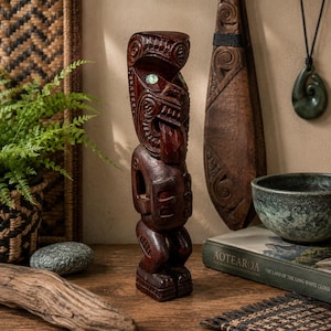 Peut inclure: Une statue tiki en bois sculpté avec des détails complexes et une finition brun rougeâtre foncé. La statue représente une forme humaine stylisée avec une tête proéminente et des traits faciaux détaillés. Une petite pierre ronde irisée est incrustée dans le front. Fait partie d'une collection d'art polynésien.