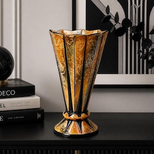May include: A decorative vase with a flared, fan-like shape. The vase features a gold and black color scheme with vertical stripes. The base is round and the top has a scalloped edge. The vase is sitting on a black surface.