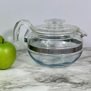 May include: A clear glass teapot with a handle and a lid. The teapot has a silver band around the middle and a spout for pouring. A green apple is visible on the left side of the image.