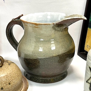 May include: A ceramic pitcher with a brown handle and a white, brown, and grey glaze. The pitcher has a wide, round body and a spout on the side.