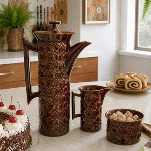 May include: A brown ceramic coffee set with a tall coffee pot, creamer, and sugar bowl. The set features a repeating circle pattern. A cake with cherries and a plate of pastries are also visible.