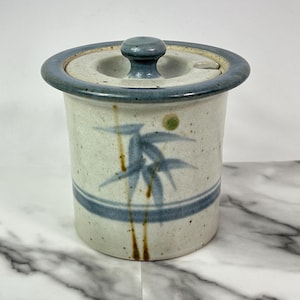 May include: A ceramic jar with a lid, featuring a blue and white bamboo design. The jar has a cylindrical shape with a blue rim and a matching knob on the lid. The bamboo design is in blue and brown.