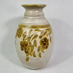 May include: Off-white ceramic vase with a floral design in shades of brown and tan. The vase has a rounded body and a narrow neck. The floral pattern is repeated around the vase.