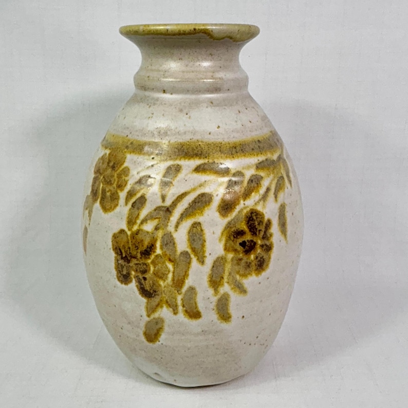 May include: Off-white ceramic vase with a floral design in shades of brown and tan. The vase has a rounded body and a narrow neck. The floral pattern is repeated around the vase.