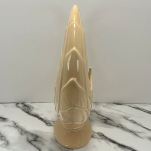 May include: A tall, beige ceramic vase with a textured, leaf-like design. The vase has a rounded base and a slender, pointed top. The surface is glossy, and the overall shape is reminiscent of a stylized plant or flower.