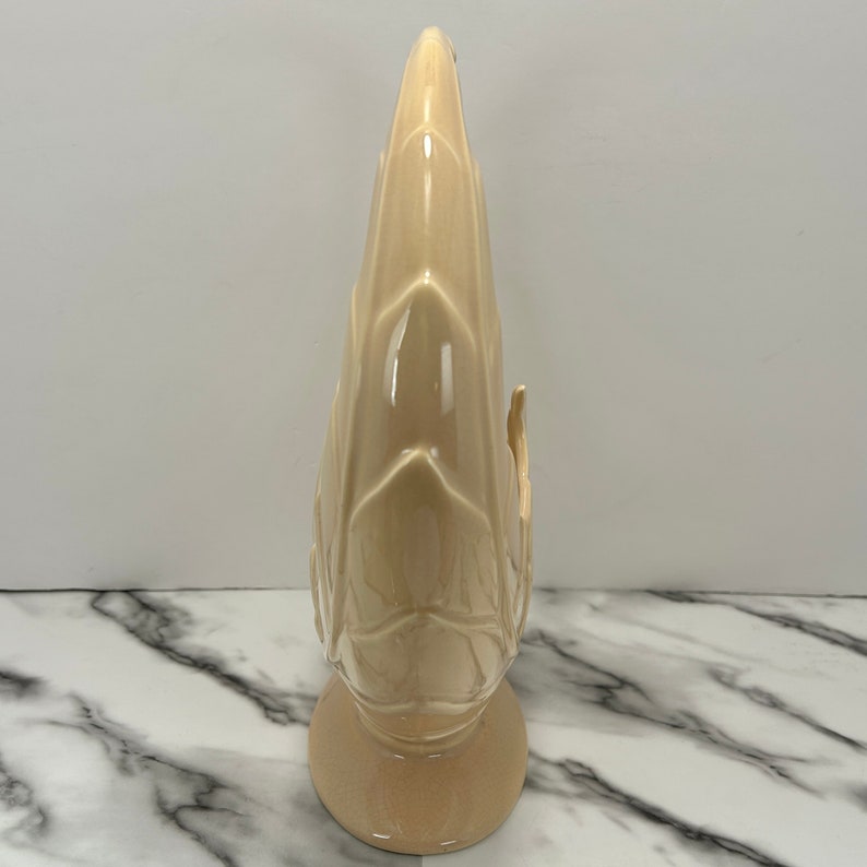 May include: A tall, beige ceramic vase with a textured, leaf-like design. The vase has a rounded base and a slender, pointed top. The surface is glossy, and the overall shape is reminiscent of a stylized plant or flower.
