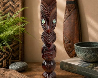 May include: A vertical dark brown wooden carving featuring a stylized figure with intricate geometric patterns and two inlaid iridescent abalone shell eyes. The sculpture is composed of stacked, rounded segments with a smooth, polished finish.