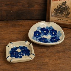 May include: Two ceramic ashtrays with blue floral patterns. One is triangular, the other square. Both have a cream base with blue painted flowers and leaves. The ashtrays are on a wooden surface.