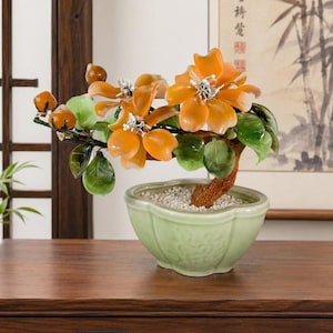 May include: A decorative bonsai tree with orange flower blossoms and green leaves. The tree is in a light green ceramic pot filled with small white stones. The bonsai is displayed on a dark wooden surface.