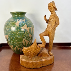 May include: A wooden figurine of a man in a hat standing on a boat. The figurine is carved in detail and has a rustic look. The man is holding a small object in his hand. The figurine is on a wooden base with the name "L. Eppensteiner" carved into it.