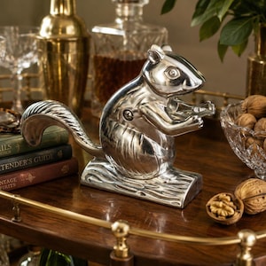 May include: A silver-toned metal nutcracker shaped like a squirrel sitting on a textured log base. The squirrel has a smooth, reflective finish with a visible screw on its side and is depicted holding its paws together in a forward-facing position.